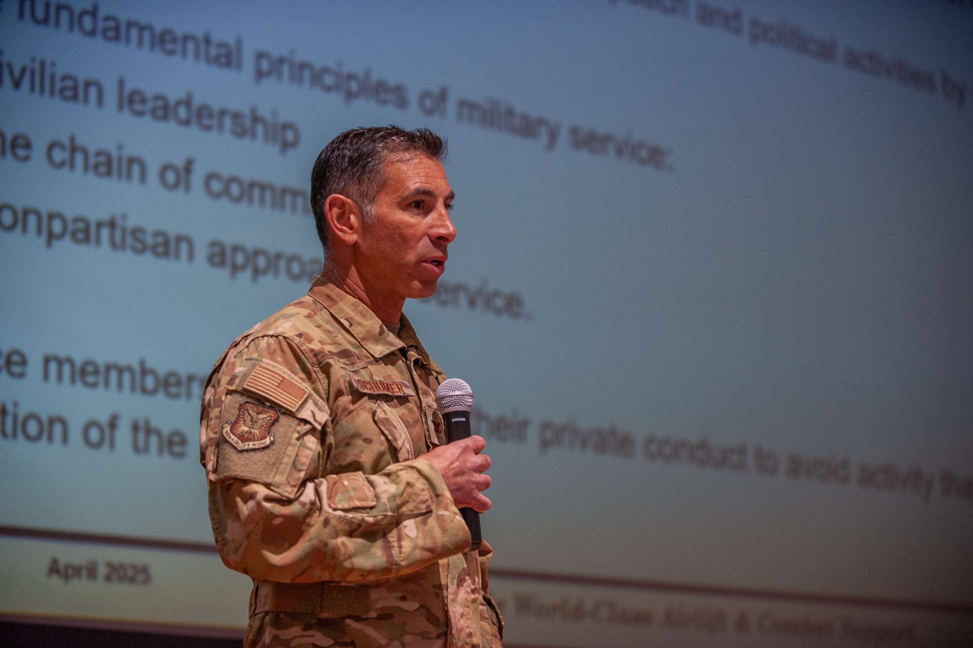A man in military uniform holding a microphone standing in front of a large presentation in the background.