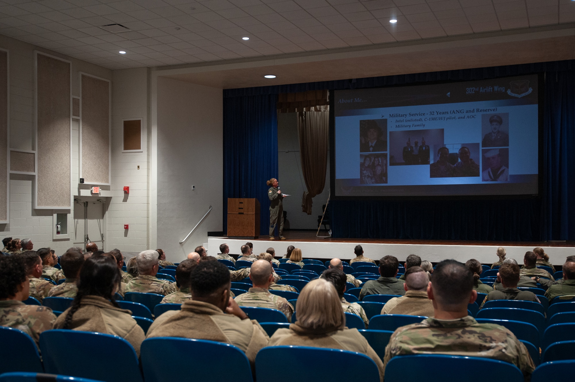 A woman in a flight suit on a theater stage in front of a crowd of seated people in military uniform.