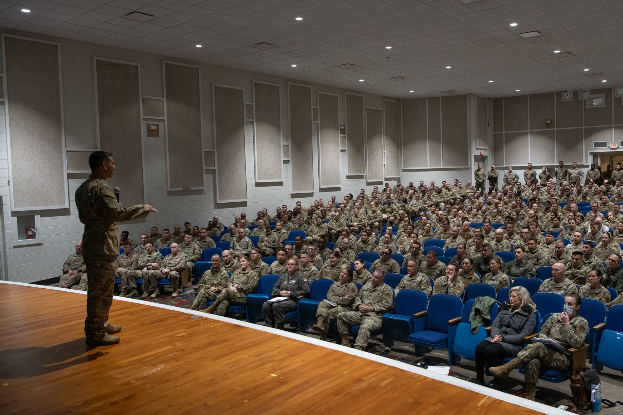 A man in military uniform on stage speaking to a crowd of military personnel in theater seating.