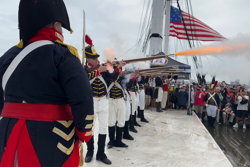 Sailors in historic garb stand in a line and simulate firing muskets while a crowd watches.