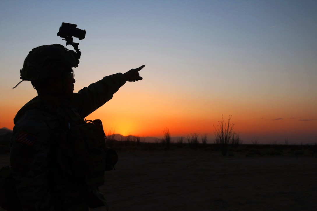 A soldier in lowlight points to an orange and blue sky.