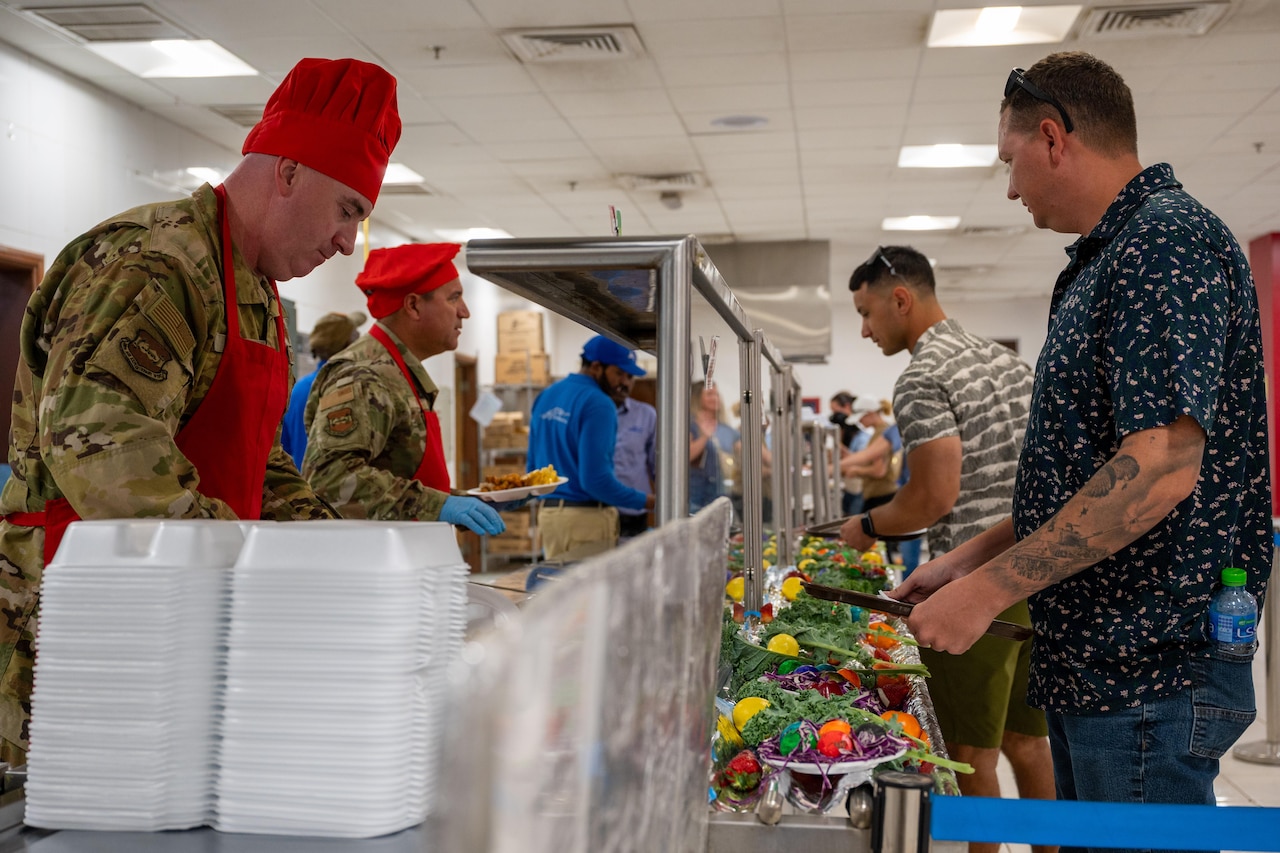 Two men in military camouflage uniforms and red chef’s hats serve food to men in casual clothing.