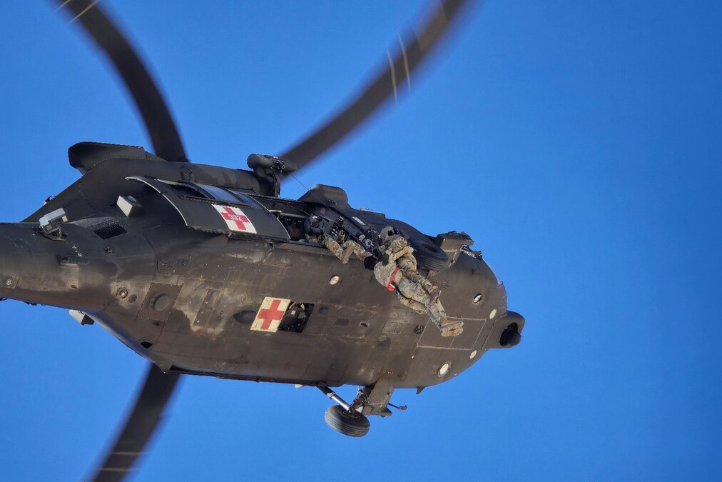 Two soldiers in harnesses hang out of a helicopter in flight and simulate a rescue.