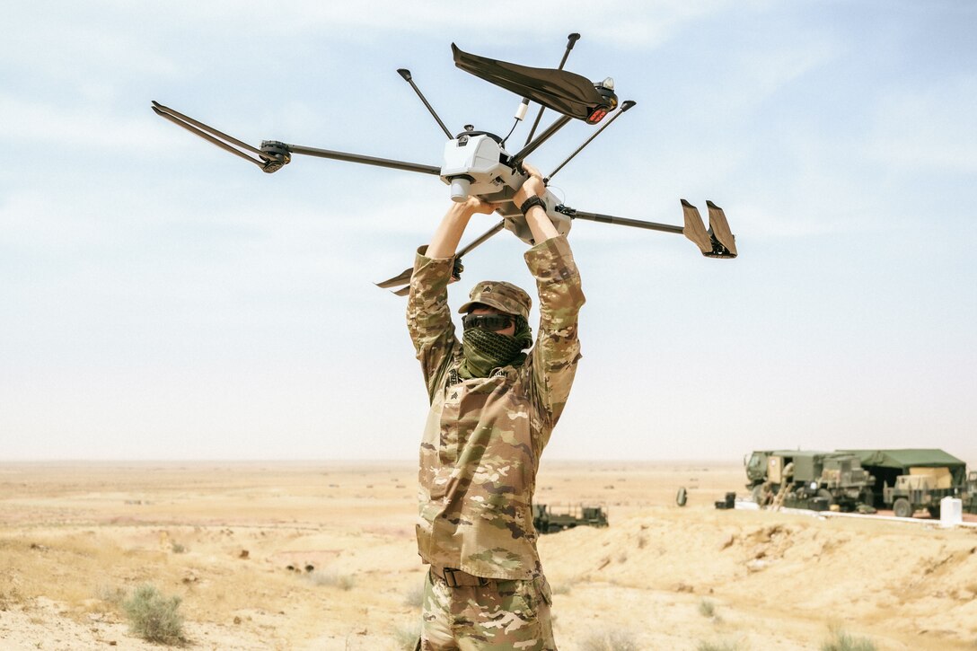 A soldier wearing a neck scarf holds up a drone while standing in a flat desert with military vehicles in the background.