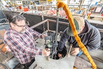 Josh Maya (left), Waterfront Support and welding work lead, Code 100TO.3, Moonshine Lab; and Bryan Hara (right), Welding Instructor, Advanced Continuous Training Development, Shop 26 lower an Encompass Welding Machine in a Rudder Bore mock-up April 9, 2025, at Puget Sound Naval Shipyard & Intermediate Maintenance Facility in Bremerton, Washington. (U.S. Navy photo by Jeb Fach)
