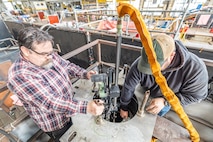 Josh Maya (left), Waterfront Support and welding work lead, Code 100TO.3, Moonshine Lab; and Bryan Hara (right), Welding Instructor, Advanced Continuous Training Development, Shop 26 lower an Encompass Welding Machine in a Rudder Bore mock-up April 9, 2025, at Puget Sound Naval Shipyard & Intermediate Maintenance Facility in Bremerton, Washington. (U.S. Navy photo by Jeb Fach)