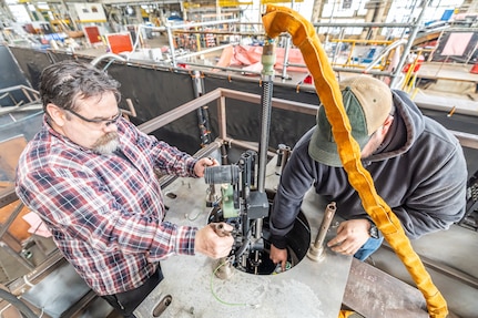 Josh Maya (left), Waterfront Support and welding work lead, Code 100TO.3, Moonshine Lab; and Bryan Hara (right), Welding Instructor, Advanced Continuous Training Development, Shop 26 lower an Encompass Welding Machine in a Rudder Bore mock-up April 9, 2025, at Puget Sound Naval Shipyard & Intermediate Maintenance Facility in Bremerton, Washington. (U.S. Navy photo by Jeb Fach)