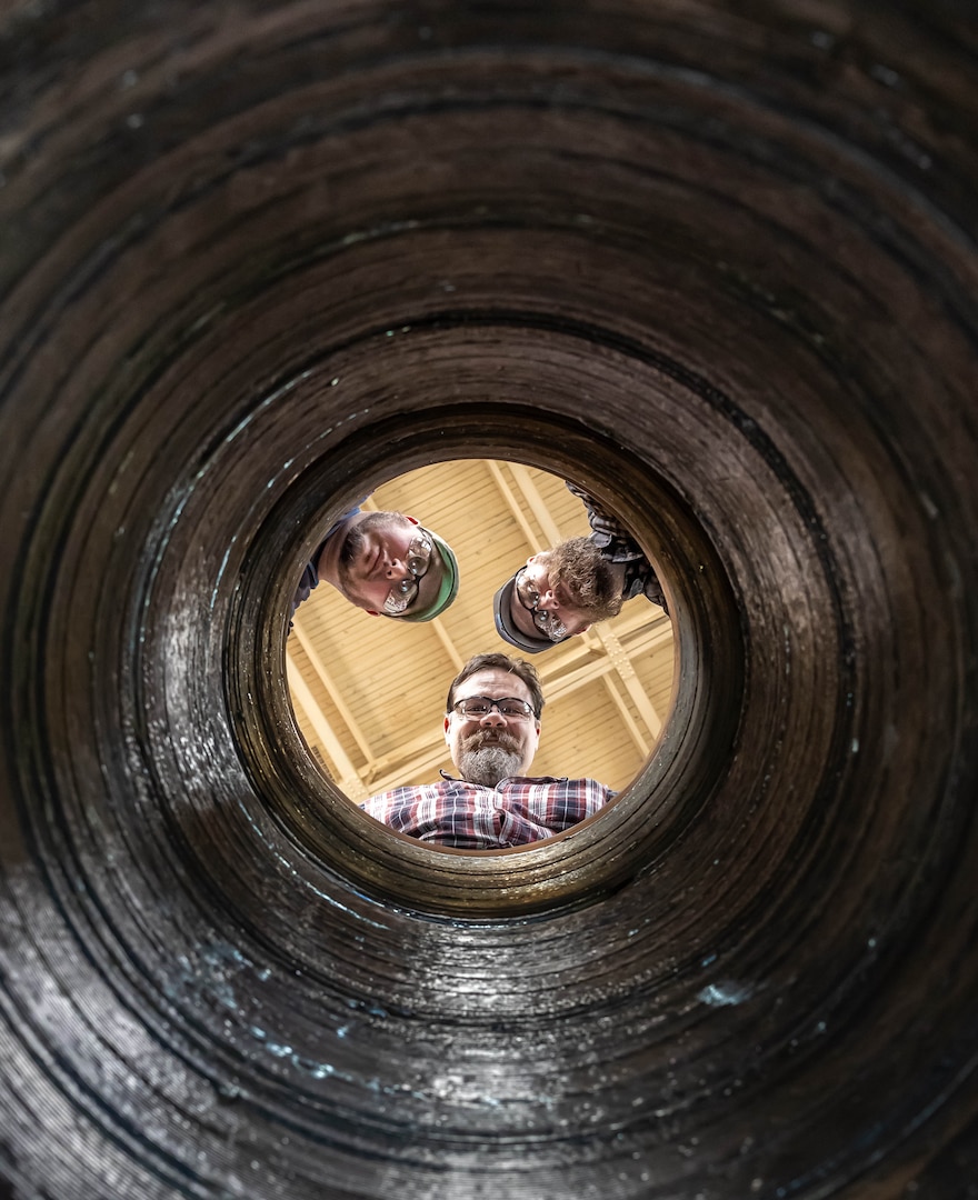 Bryan Hara (top left), welding instructor, Advanced Continuous Training Development, Shop 26; Josh Maya (center), Waterfront Support and welding work lead, Code 100TO.3, Moonshine Lab; and Josh Hughes (top right), welder training leader, Shop 26, pose for a photograph at the top of a rudder bore mockup in Building 460  April 9, 2025, at Puget Sound Naval Shipyard & Intermediate Maintenance Facility in Bremerton, Washington. The mock-up is used for training welders in rudder bore repair. (U.S. Navy photo by Jeb Fach)