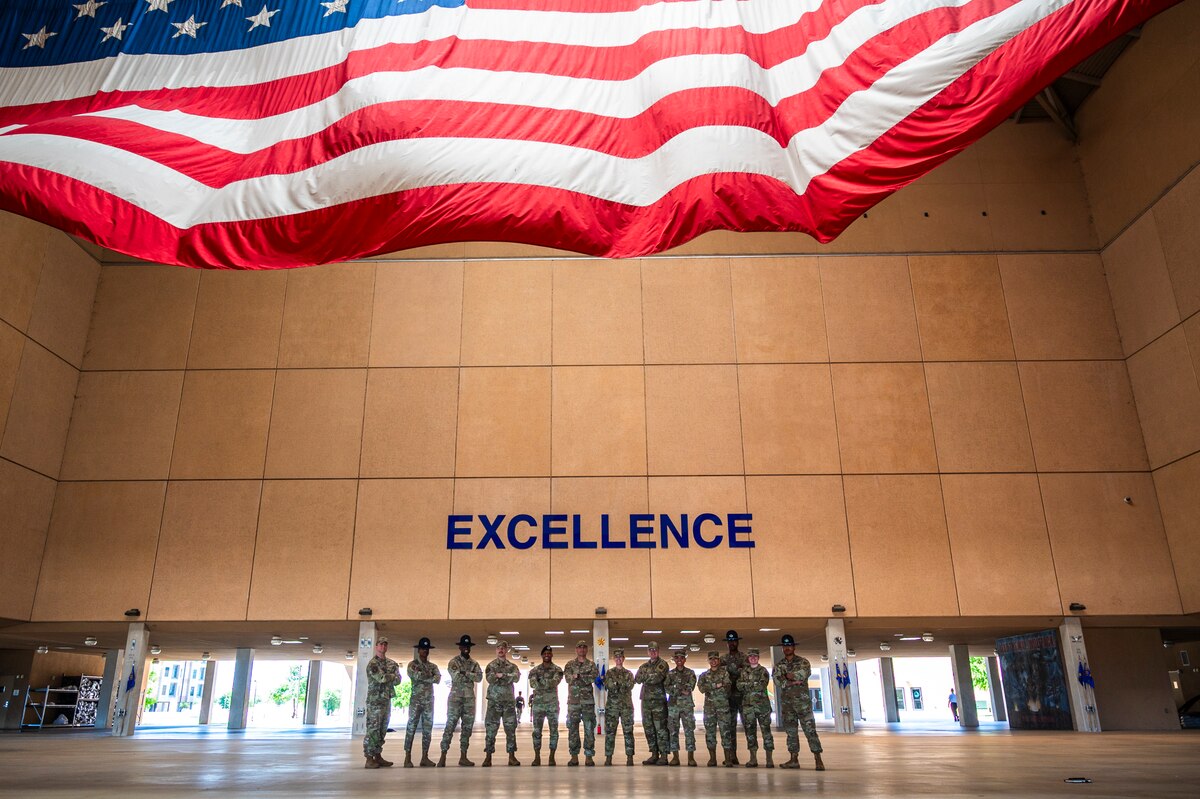 U.S. Air Force Col. Sergio Anaya, 62d Airlift Wing commander, Chief Master Sgt. Kristy Korchak-Campbell, 62d AW command chief, a group of Team McChord NCOs, and several 331st Training Squadron military training instructors stand under the American flag in the Peterson Training Complex atrium at Joint Base San Antonio-Lackland, Texas, April 9, 2025.