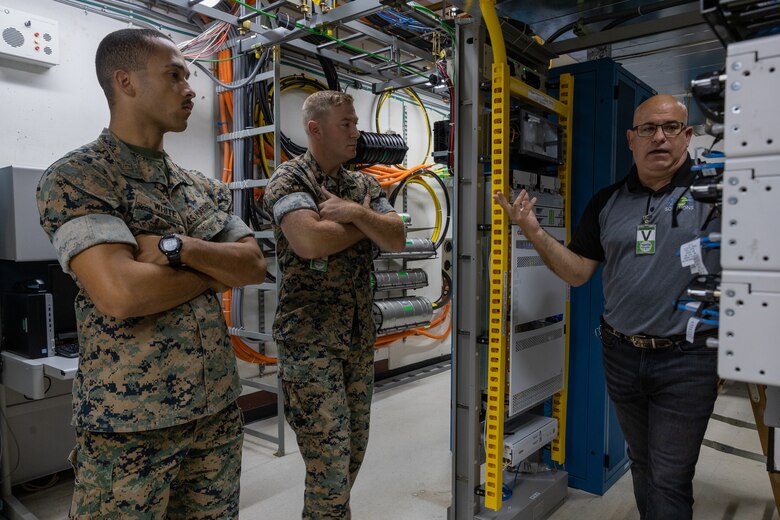 Two Marines strand with arms crossed as a contractor technician explains work going on with a telecomminication system.