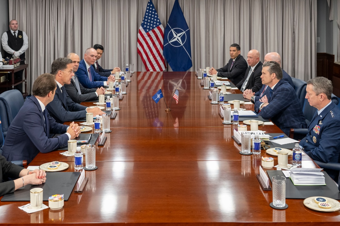 Government officials sit around a rectangular table with suits and flags at the end of the room.