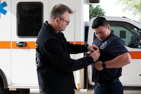 A civilian Air Force paramedic pins an EMS Field Save Pin onto the shirt of a Senior Airman paramedic in front of an ambulance