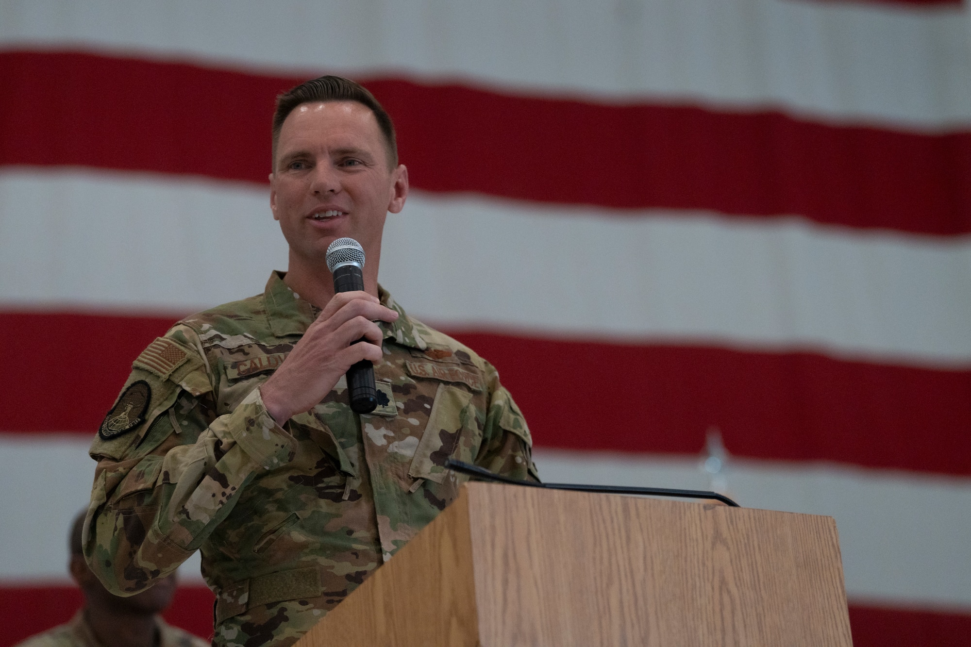 An airman stands at a podium with a microphone with the American flag in the background.