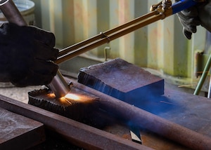 a hand comes in to frame from the left holding a sliver rod it is hitting what looks like rusted metal plate on a metal table causing sparks. A torch, with the tip being in the sparks, is being held by hands coming in from the other side of the frame.