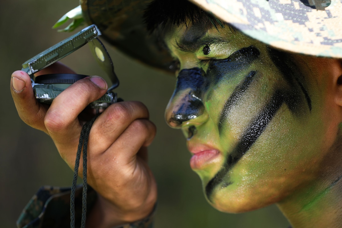 A Marine wearing camouflage face paint and a cover looks through a green compass.