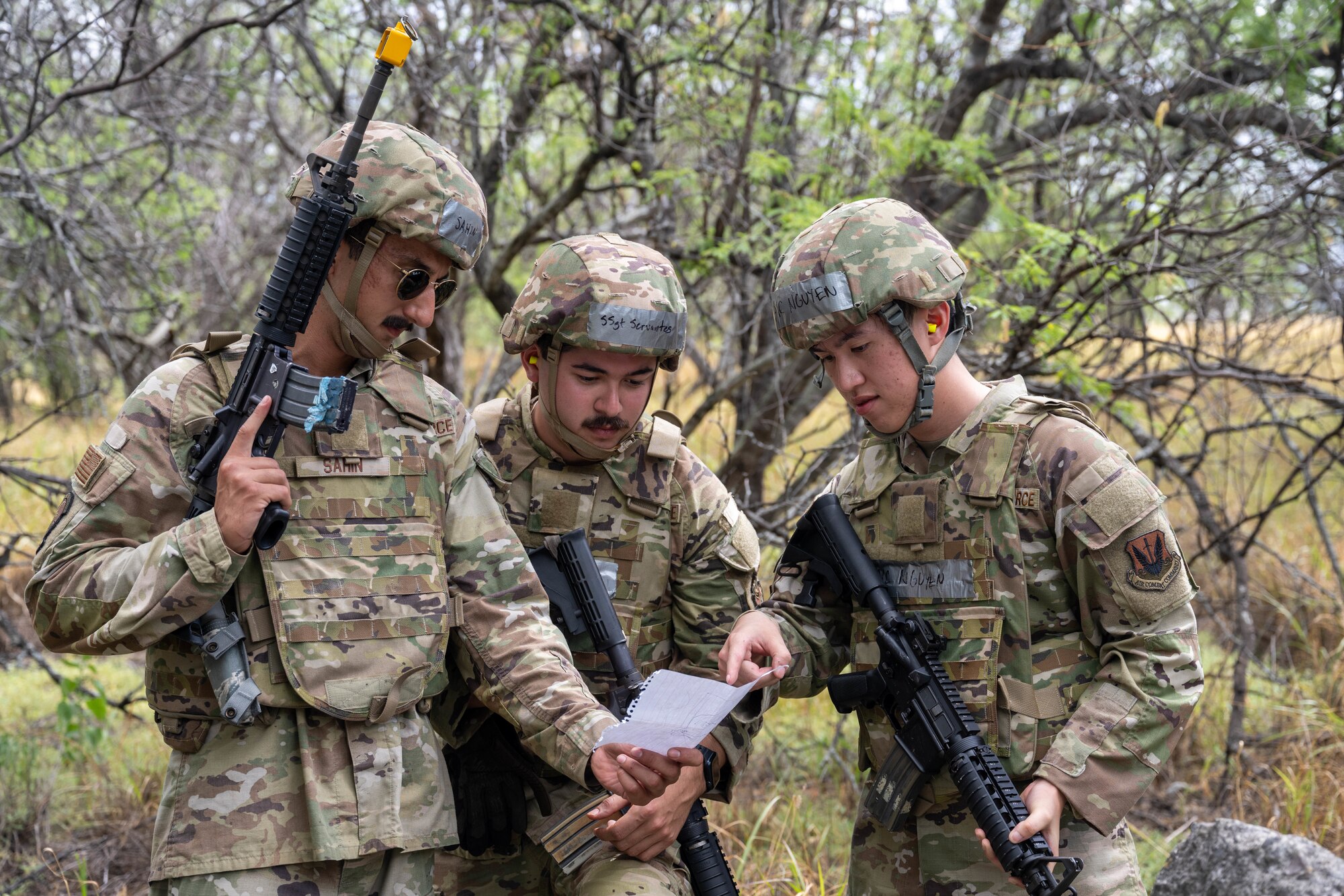 Three U.S. Air Force Airmen look at their notes while holding their M4 assault rifles during a training session in the woods.