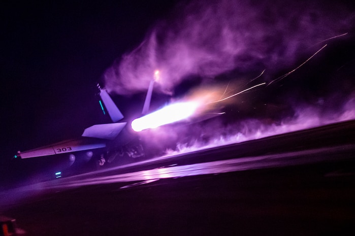 A military aircraft takes off from a ship, leaving purple smoke behind against a dark sky.