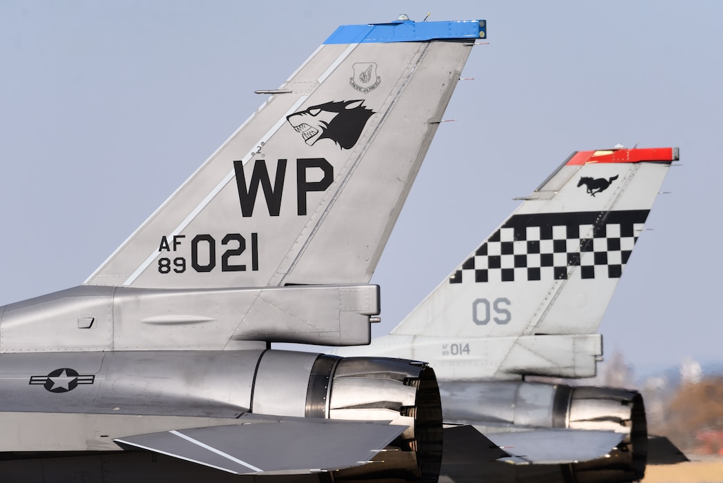 Two F-16 tails are shown side-by-side, one with a 8th Fighter Wing "Wolf Pack" logo and one with a 51st Fighter Wing "Mustang" logo.