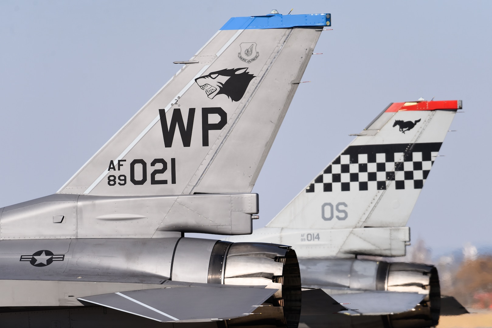 Two F-16 tails are shown side-by-side, one with a 8th Fighter Wing "Wolf Pack" logo and one with a 51st Fighter Wing "Mustang" logo.