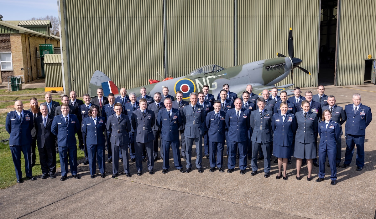 uniformed U.S., U.K., an Australian military members stand in front of Battle of Britian Memorial Flight hangar