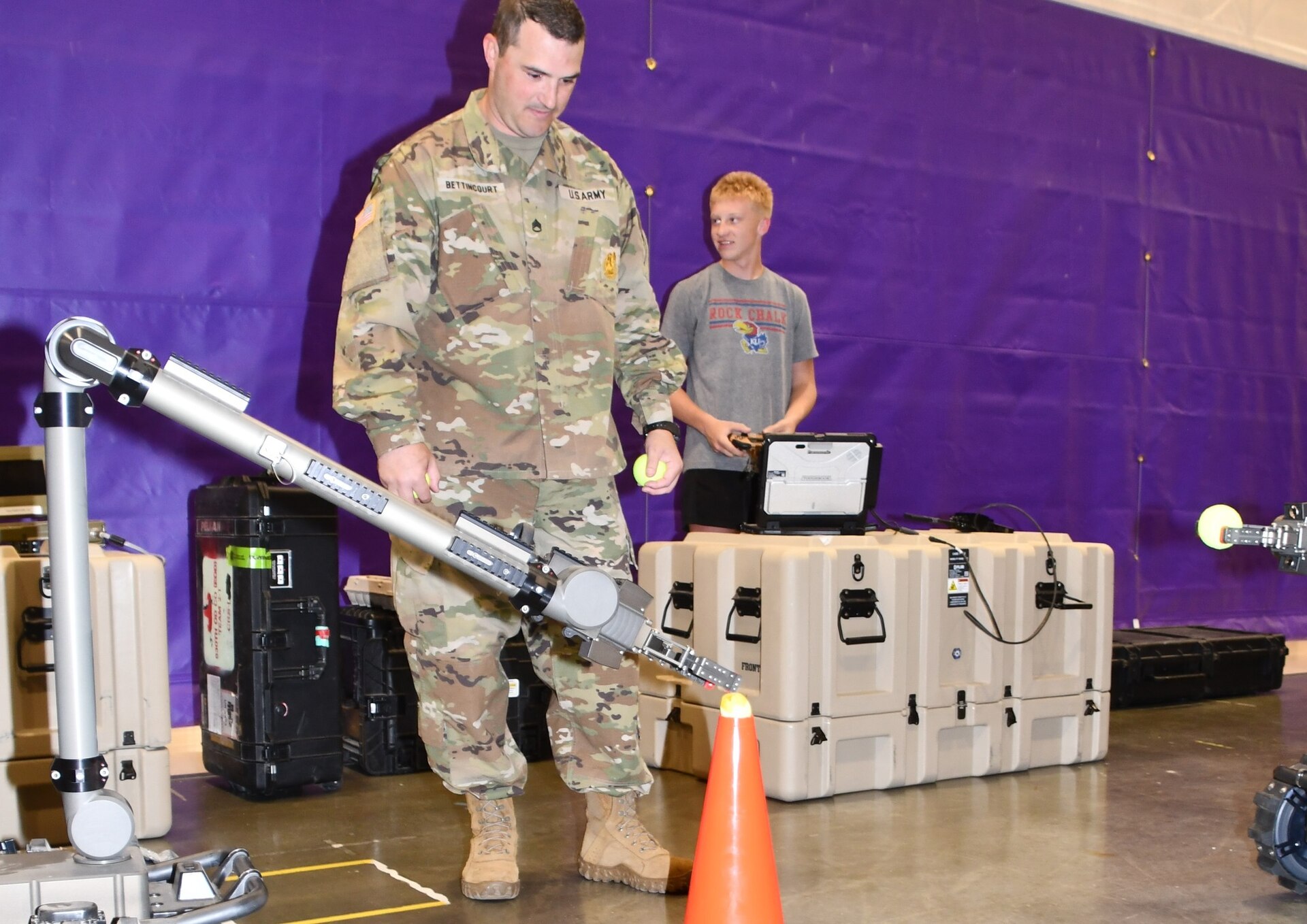 Staff Sergeant Bettincourt, Overland Park Army Recruiting Station commander, demonstrates the capabilities of a robotic arm used in Explosive Ordnance Disposal (EOD) while a student operates the controls during a STEM engagement event at Spring Hill High School in Spring Hill, Kansas. The event introduced students to Army technology and its real-world applications in critical missions.