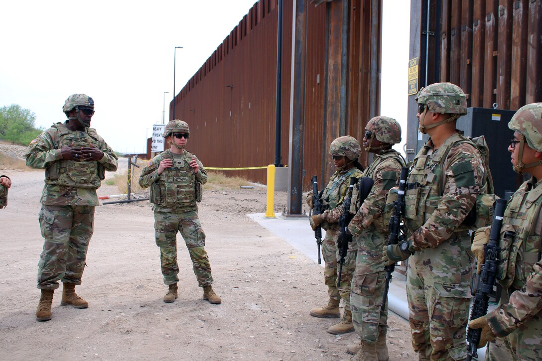Soldiers carrying weapons gather for a briefing near a large fence on a gloomy day.