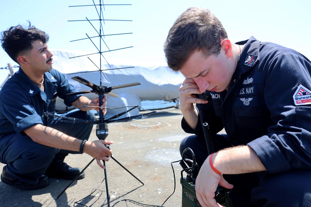 Two sailors kneel while setting up an antenna against a blue sky.