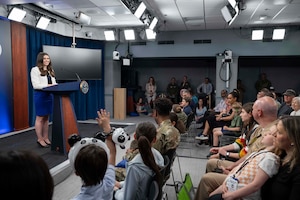 A woman in business attire stands behind a lectern. Seated in front of her is a room full of adults and children.