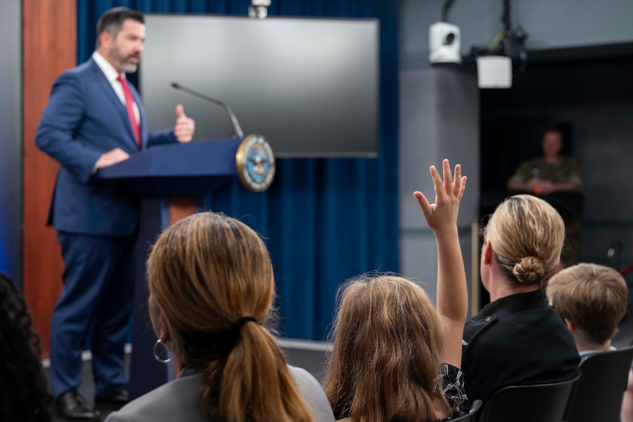 A man wearing business attire stands behind a lectern. A seated child raises their hand.