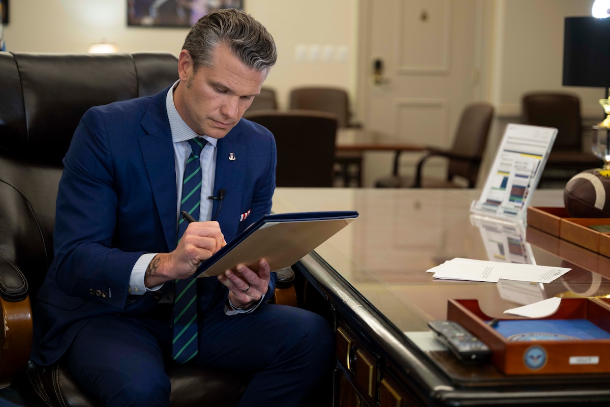 A man in a suit sits at a polished wooden desk in an office, signing a document with a pen.