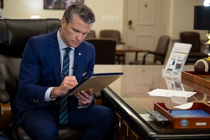 A man in a suit sits at a polished wooden desk in an office, signing a document with a pen.