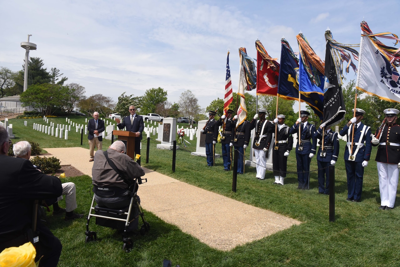 A man standing in a cemetery speaks to a seated crowd.