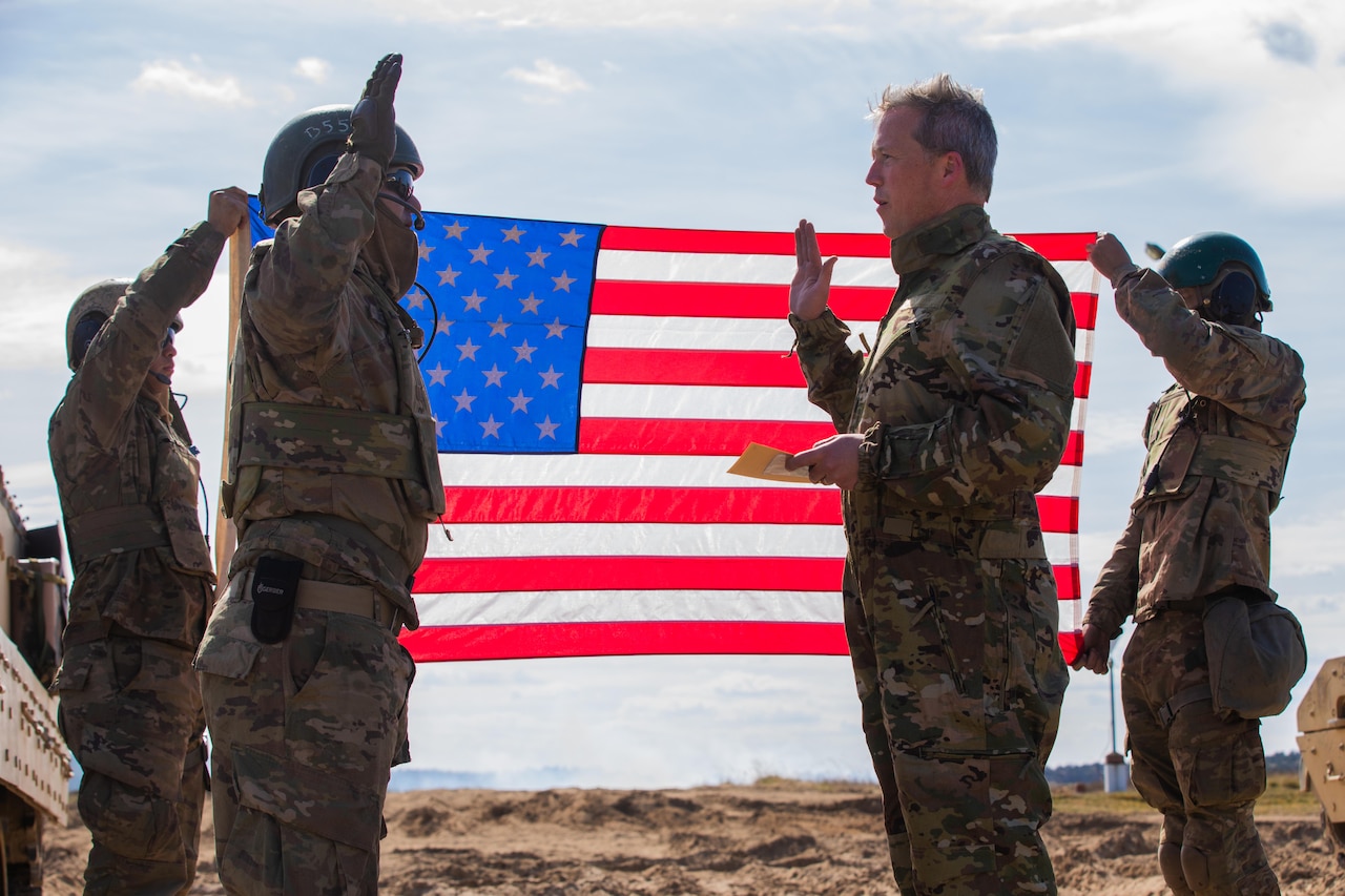 Two men in camouflage uniforms, with their right hands raised in the air, stand in front of an American flag.