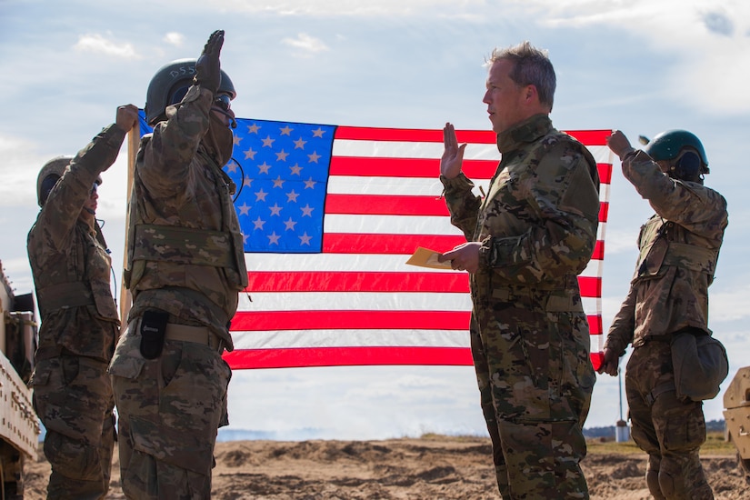 Two men in camouflage uniforms, with their right hands raised in the air, stand in front of an American flag.