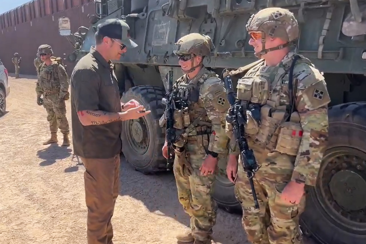 A man speaks with two soldiers in front of a combat vehicle.