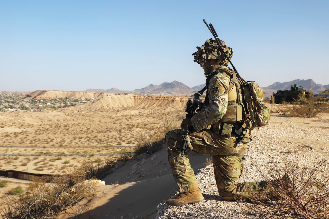 A soldier takes a knee outdoors in a large mountainous area.