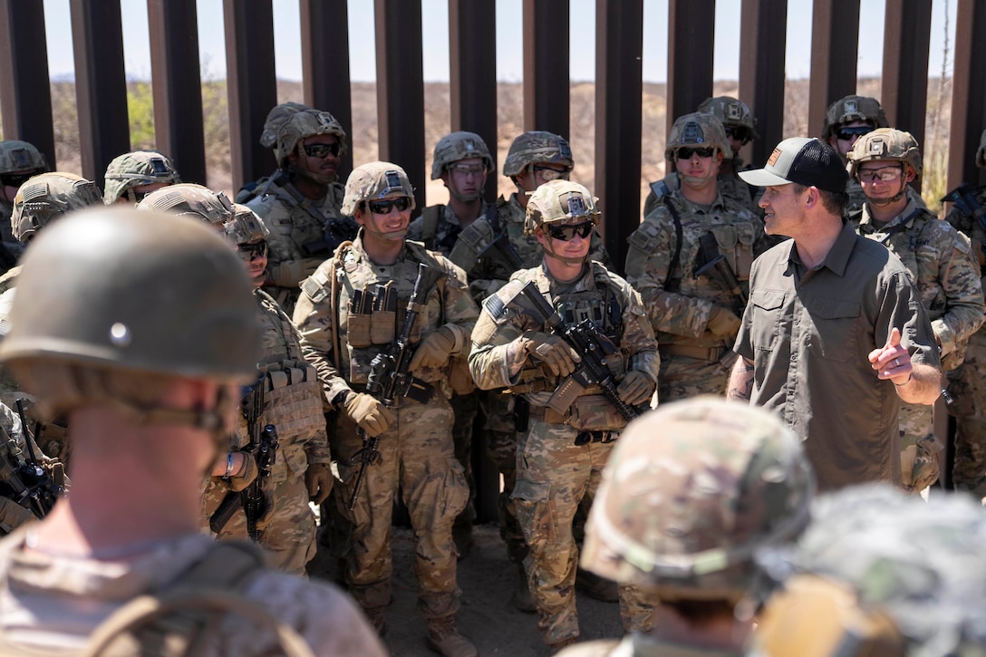 Defense Secretary Pete Hegseth talks with a group of service members surrounding him with the southern border wall in the background.