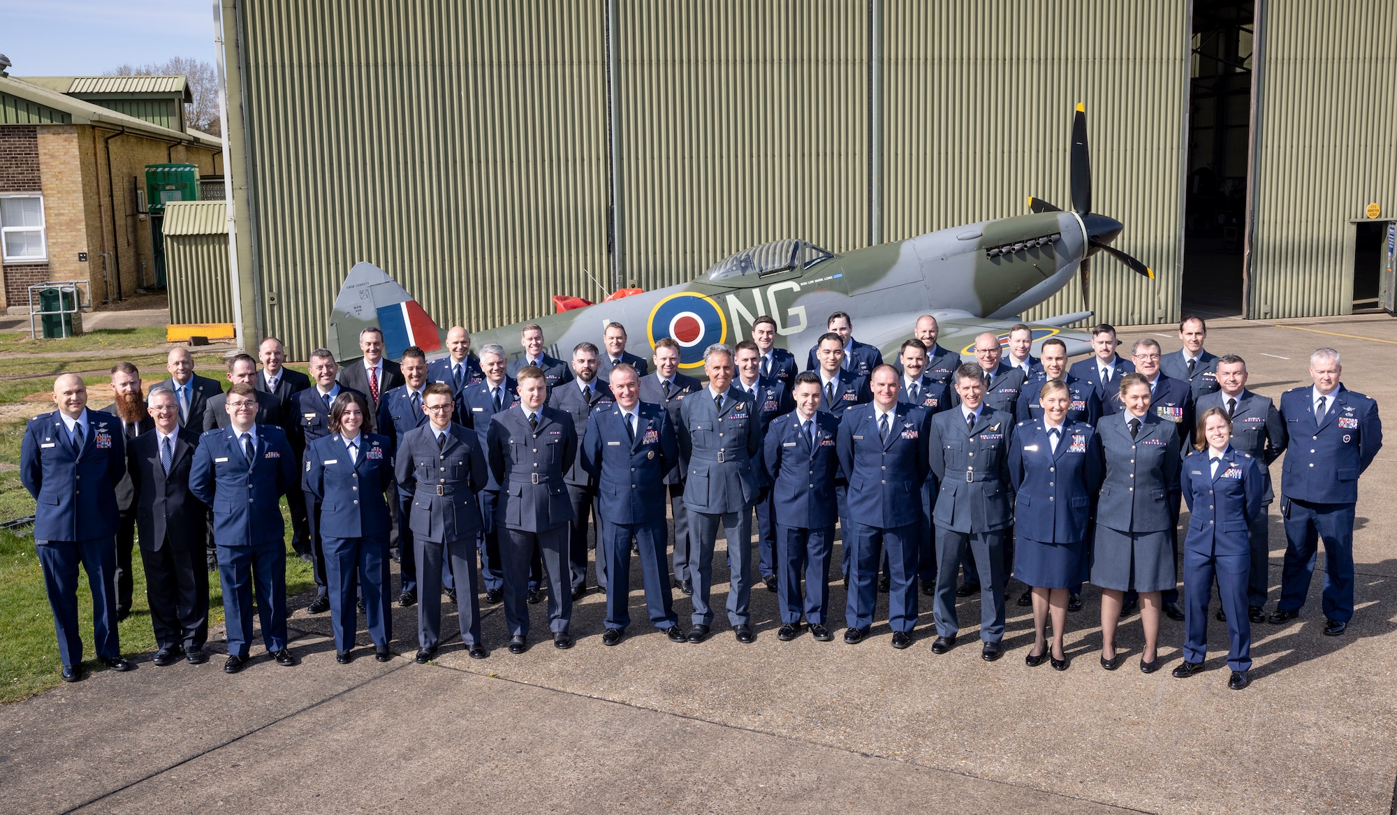 uniformed U.S., U.K., an Australian military members stand in front of Battle of Britian Memorial Flight hangar
