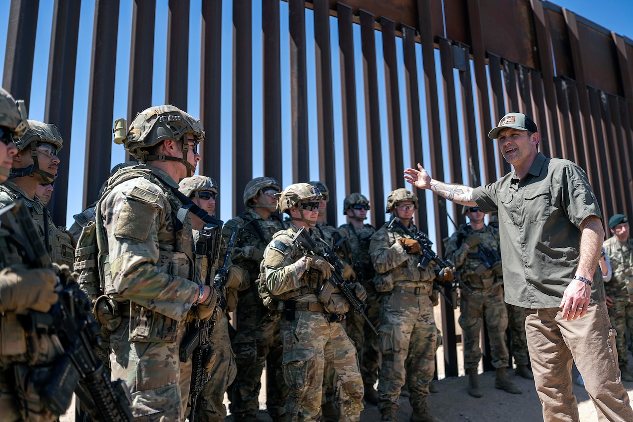 A man in civilian clothes stands in front of a dozen individuals in military uniforms and a large barricade.
