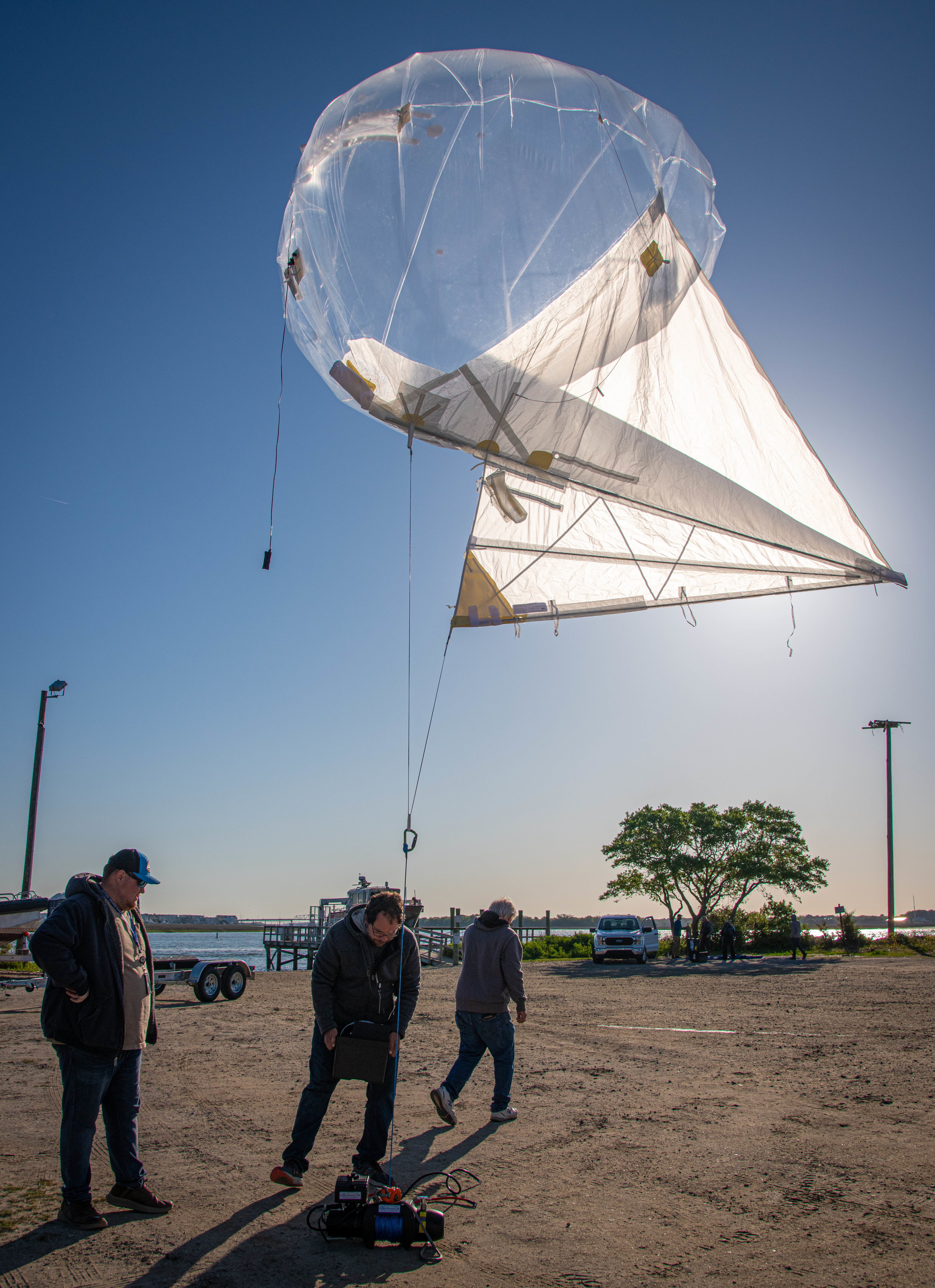 Two of three men examining a winch on the ground attached to a balloon.  One man is walking away.
