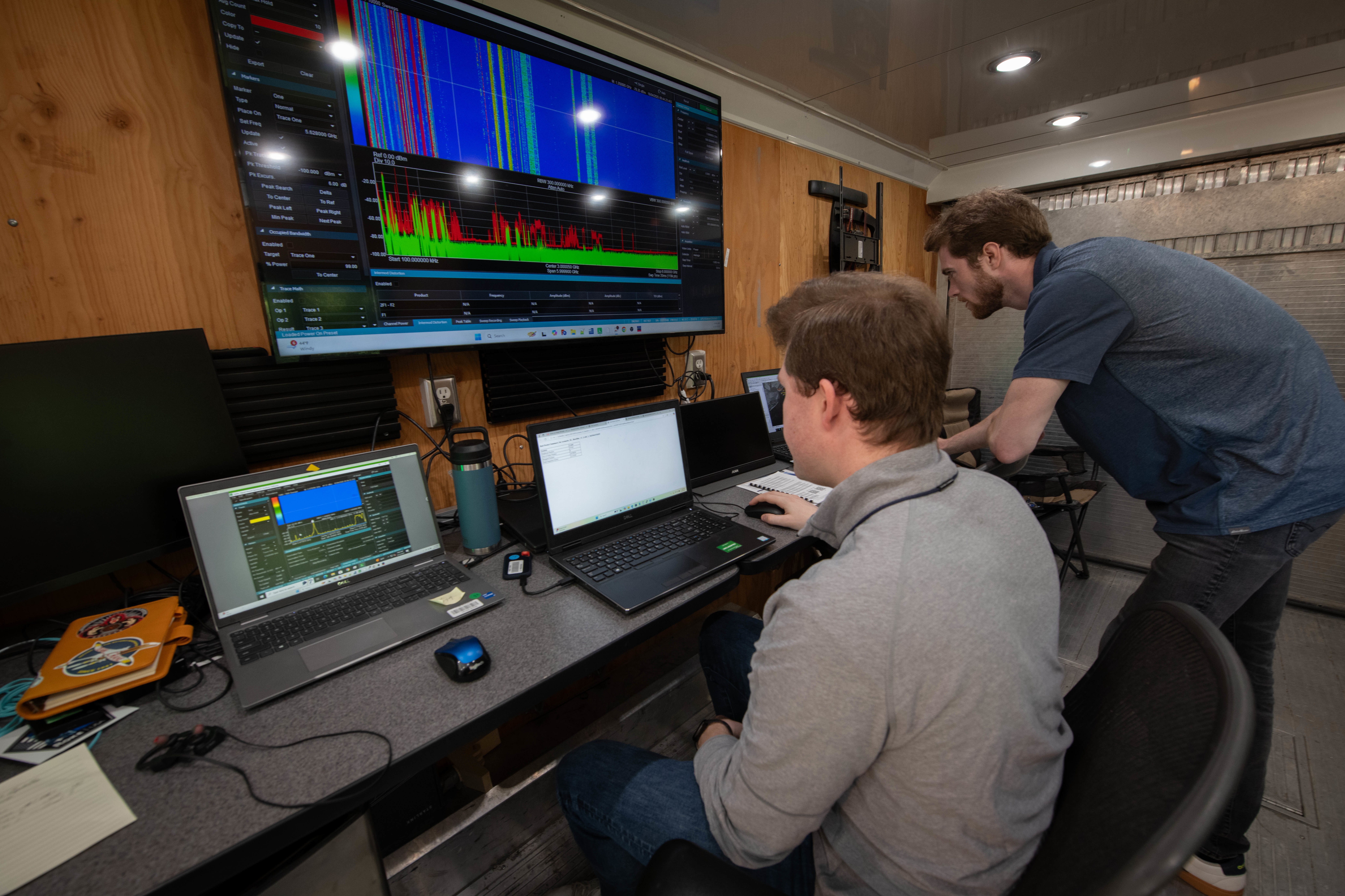 Two men looking over laptops and monitors with charts on a larger screen above.