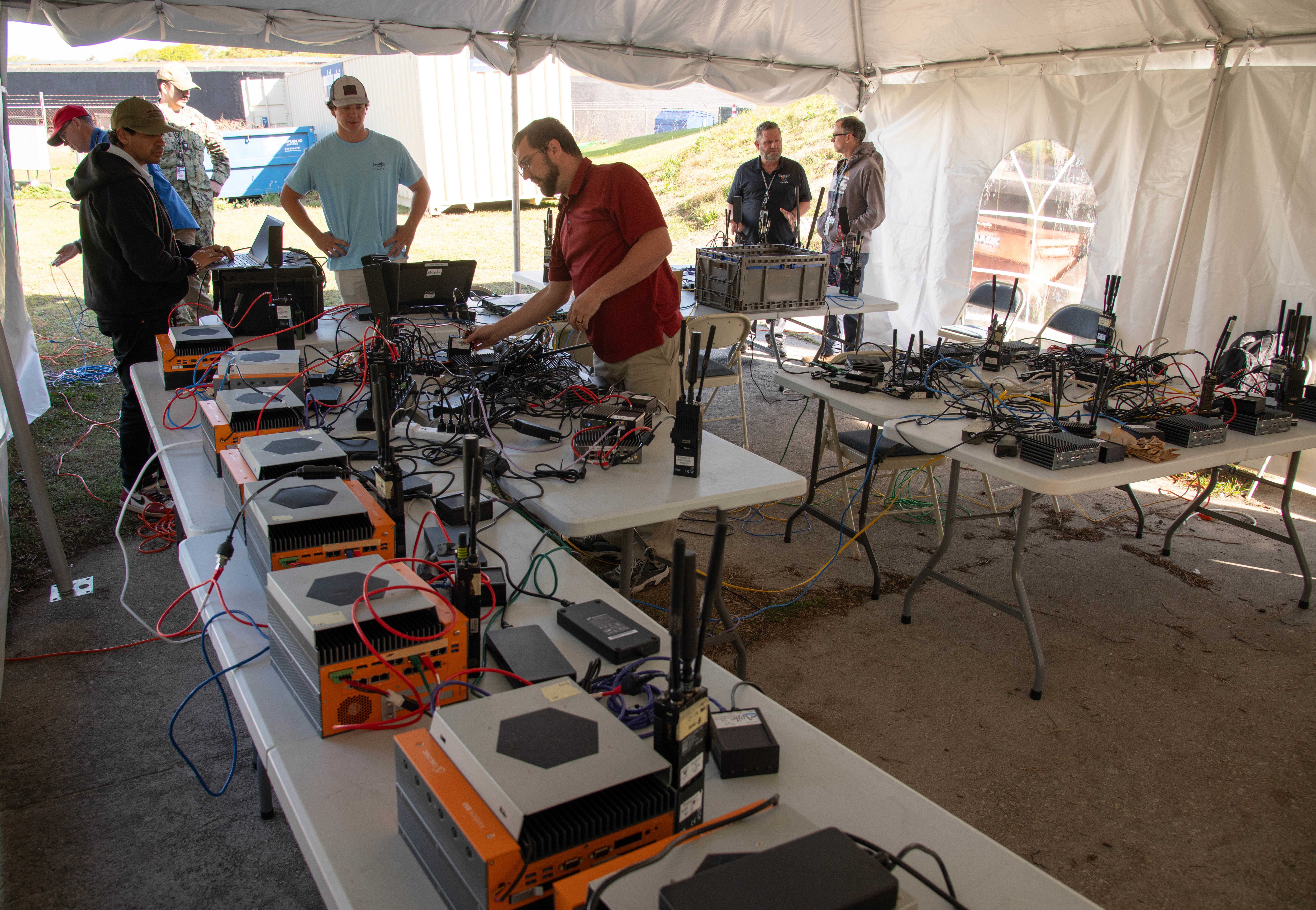 7 men looking over radio equipment on tables under a large white tent.