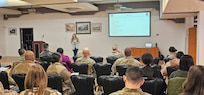 Female speaking to a seated audience of military and civilian personnel.