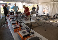 7 men looking over radio equipment on tables under a large white tent.
