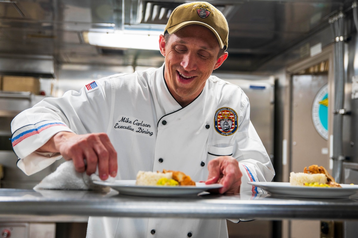 A person wearing a hat and a chef's coat smiles and wipes a plate that has food on it with a cloth.