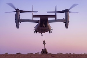 An airman slides down a rope attached to an aircraft as several other airmen kneel on the ground.