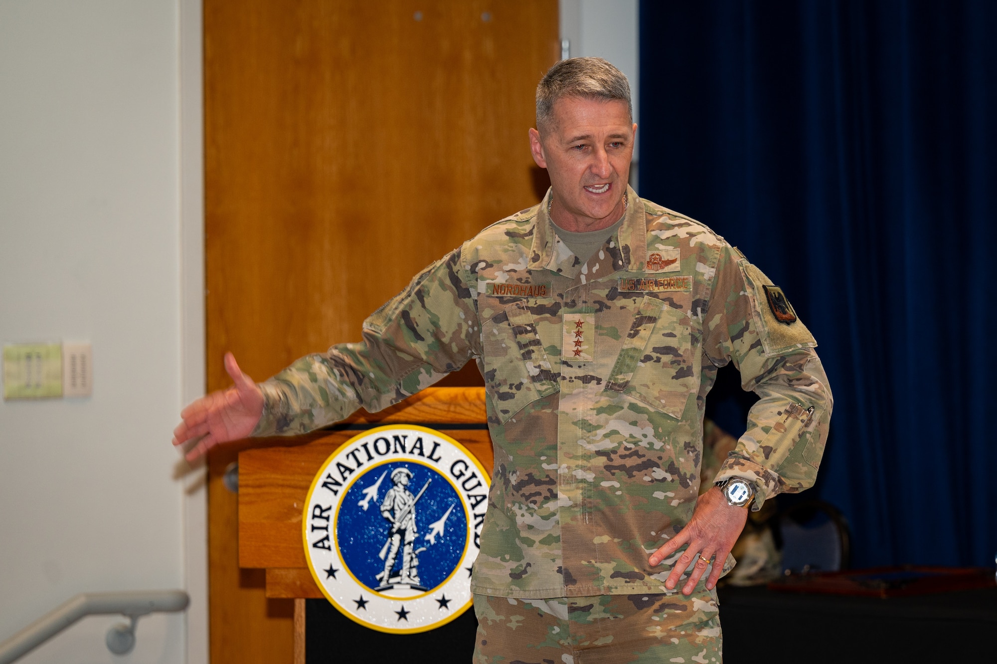A uniformed service member is seen speaking with his right hand gestured away from him and the other placed on his hip.