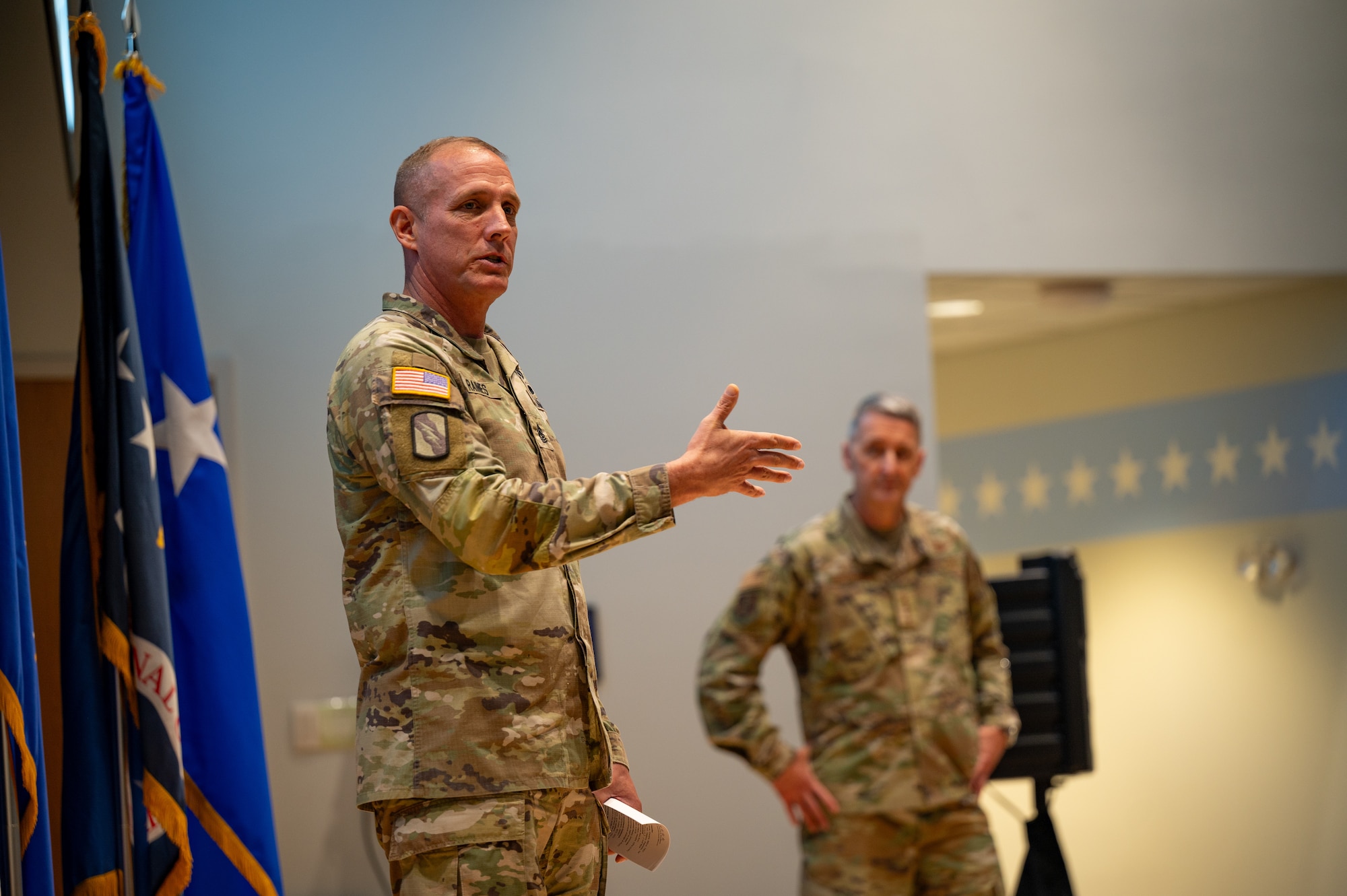 Two uniformed service members are seen from a low perspective on stage. The subject on on left is speaking while gesturing with his left hand and hold a piece of paper in his right.