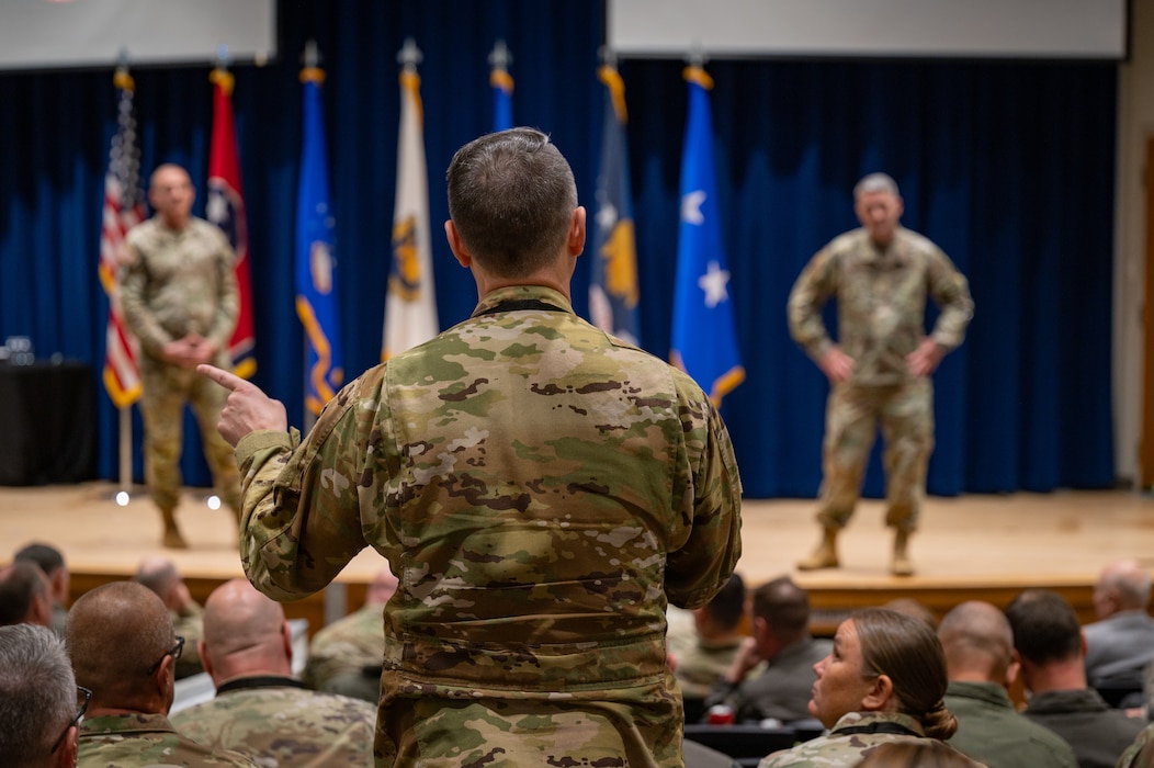 A uniformed service member is seen from behind standing up in a crowd of seated people. The subject is asking a question to two uniformed service members on a stage seen on either side of him.