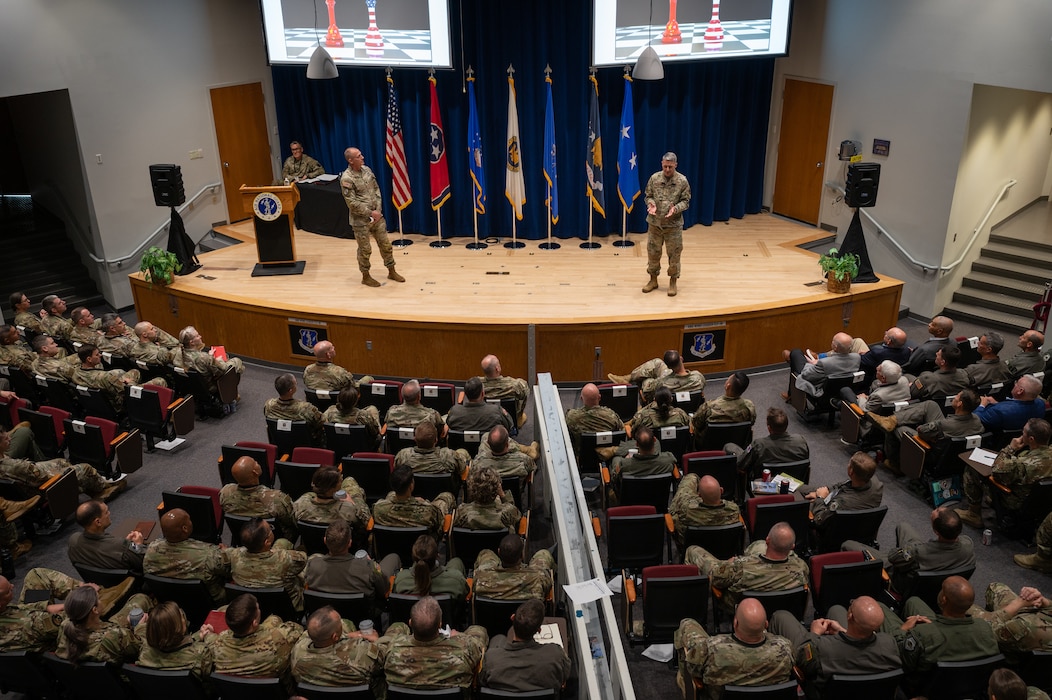 Two uniformed service members stand on stage in front of an audience of other uniformed service members in an auditorium.